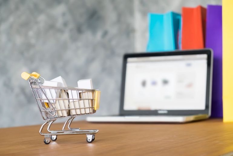 A miniature shopping cart filled with boxes in front of a laptop and some shopping bags