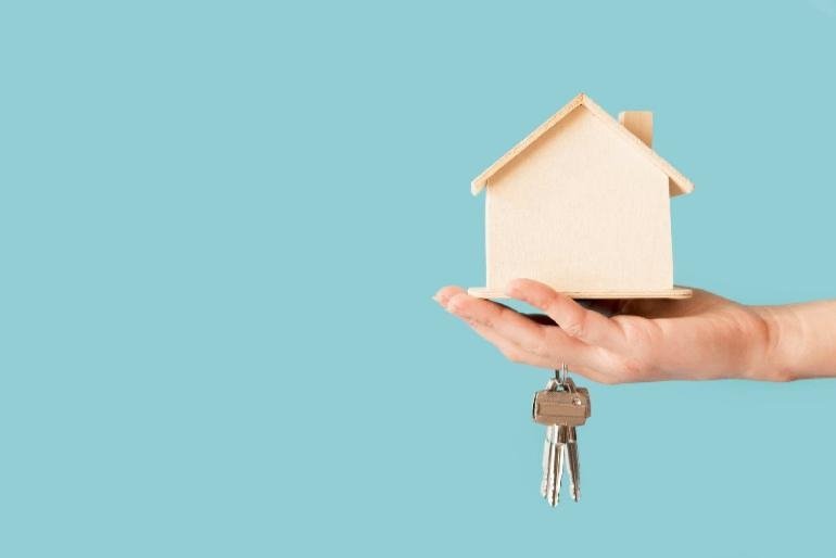 Person holding a wooden model house in the palm of their hand with house keys hanging from a finger