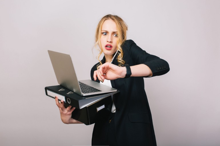 Business woman struggling to hold a laptop, folder while looking at her watch