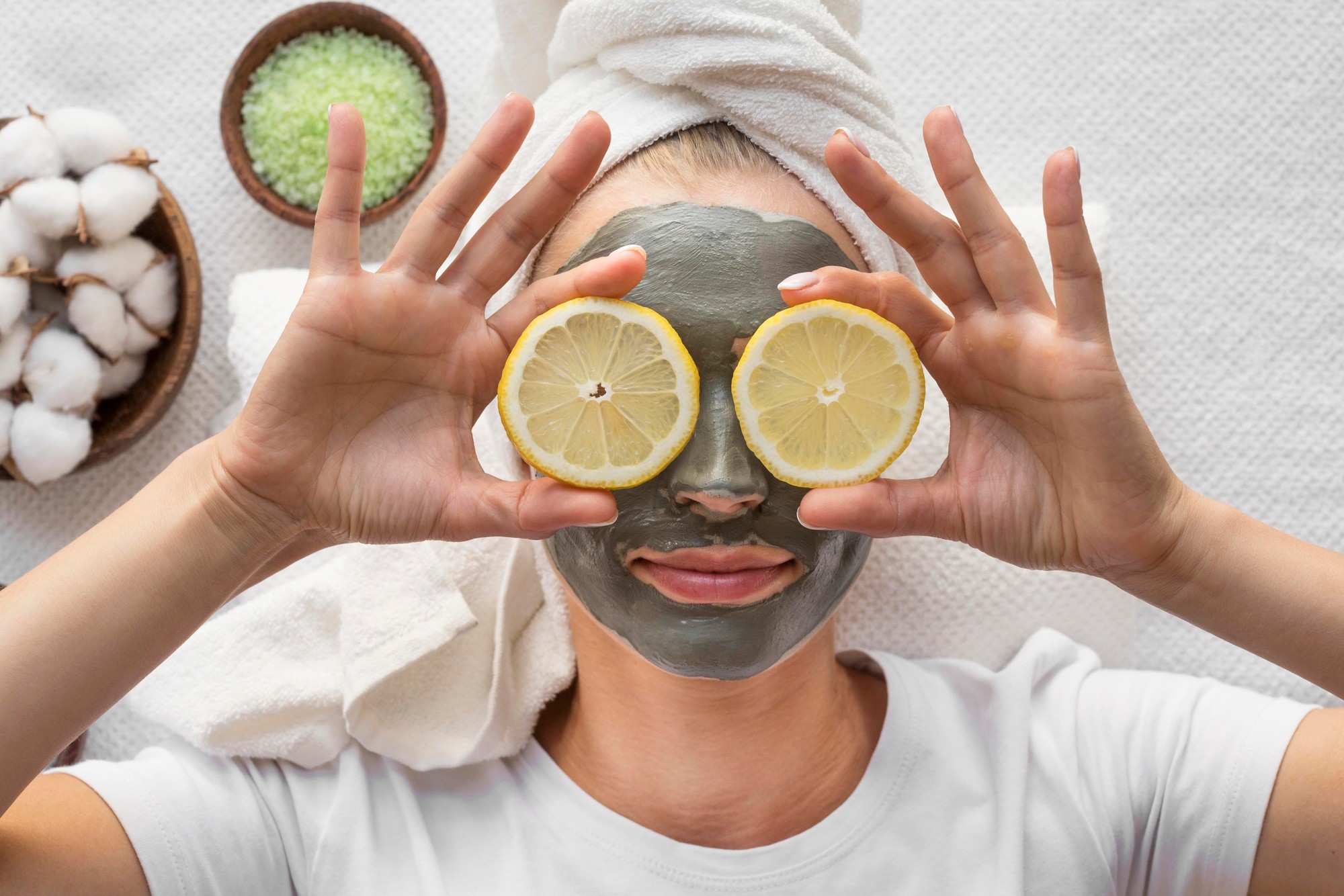 Person with a towel wrapping their hair, wearing a face mask and covering their eyes with lemons