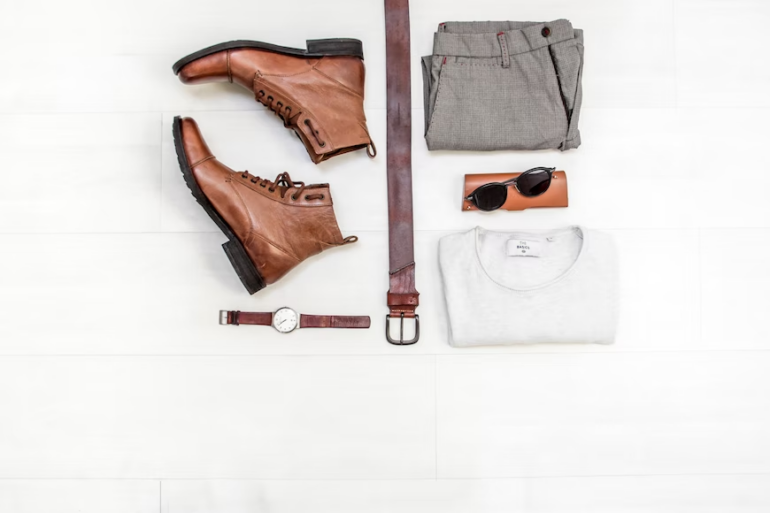 Top-down view of men's brown leather boots and belt, watch, trousers, sunglasses and white tee-shirt