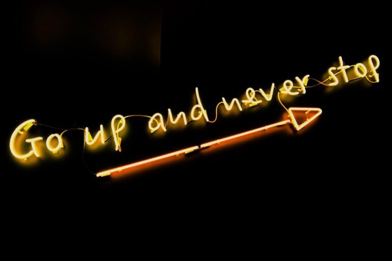 Yellow neon writing on a black background. The writing says "Go Up And Never Stop" with an arrow pointing upwards