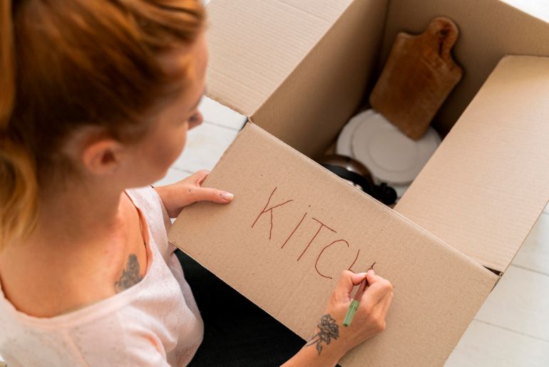 Photo of a person writing the word "Kitchen" on a carboard box that contains kitchen equipment