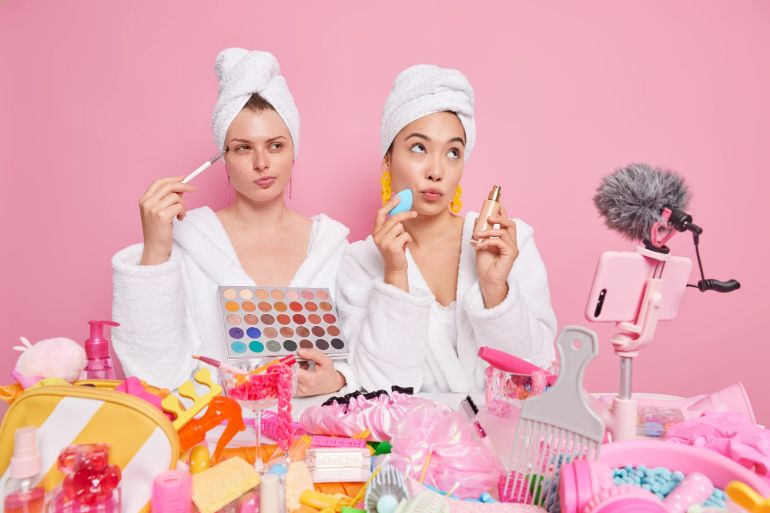 Two women wearing white dressing gowns, towels on their heads and they are surrounded by make up