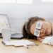 Woman asleep on a desk with fake cartoon eyes drawn on post-it notes