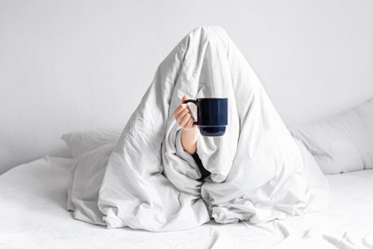 Person sitting on a sofa, hidden under a duvet holding a mug of coffee
