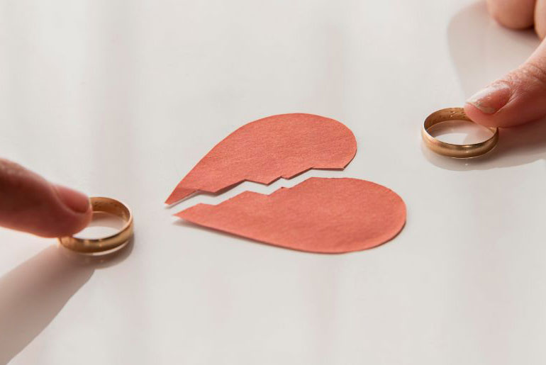A broken red paper heart with two people pushing wedding bands towards it with their fingers
