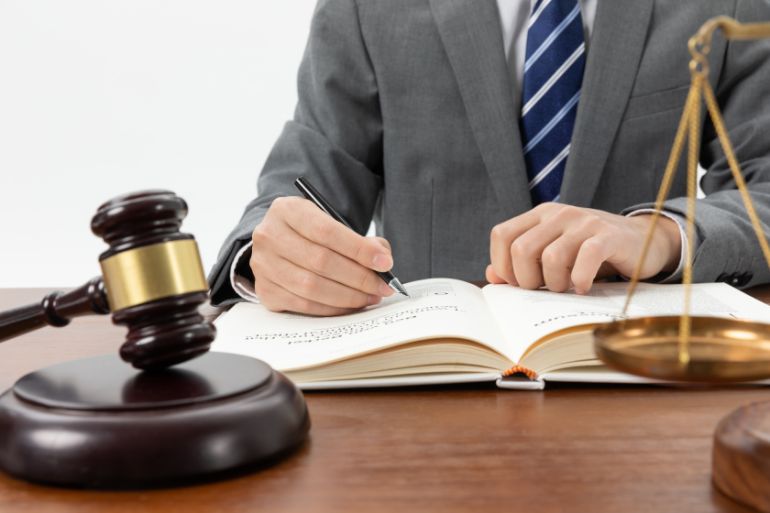 Person in a suit, writing in a large book with a gavel and scales of justice
